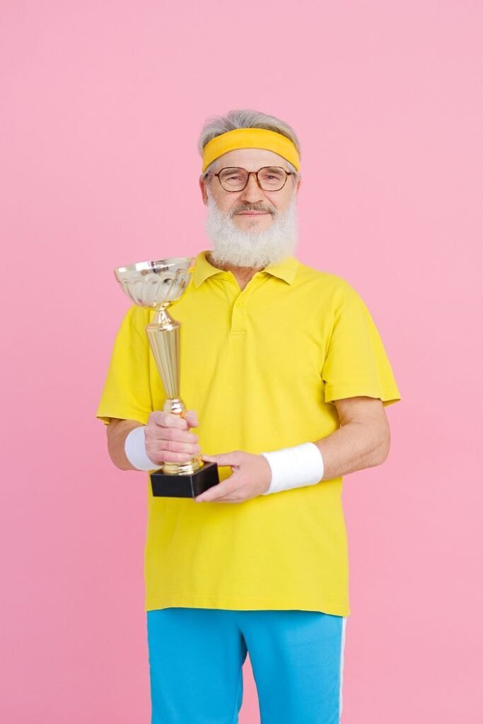Senior man in sports attire proudly holding a trophy against a vibrant pink background.